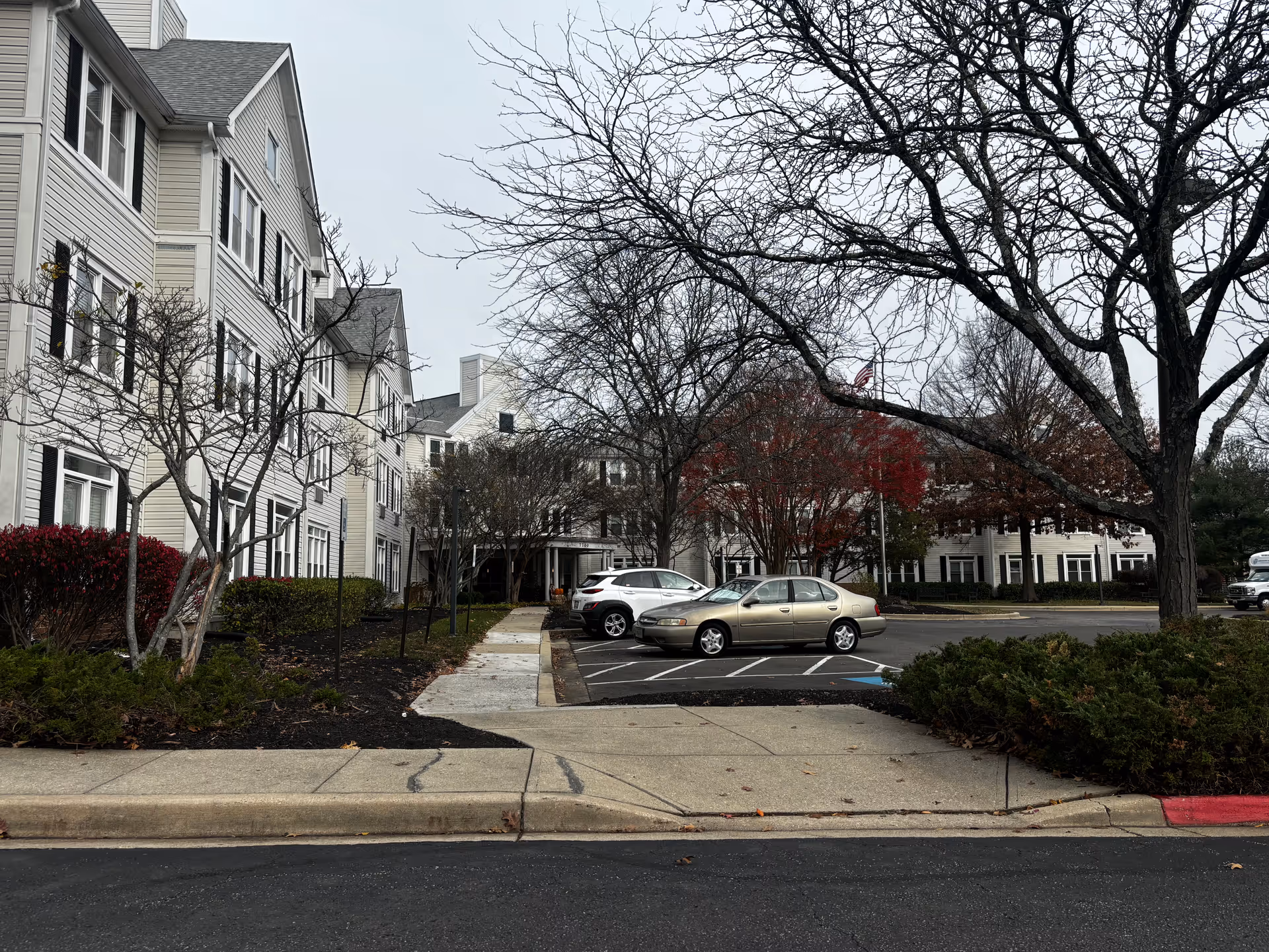 Front exterior of a multi-story beige assisted living building with a parking lot, parked cars, leafless trees, and a sidewalk.
