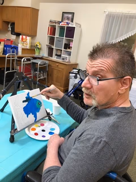 A man wearing glasses is seated at a table painting a blue fish on a canvas. The table is covered with a turquoise tablecloth and has a paint palette with various colors. Behind him, there are shelves with storage bins and some mobility aids like walkers in a room with wooden cabinets and a window with a lace curtain.