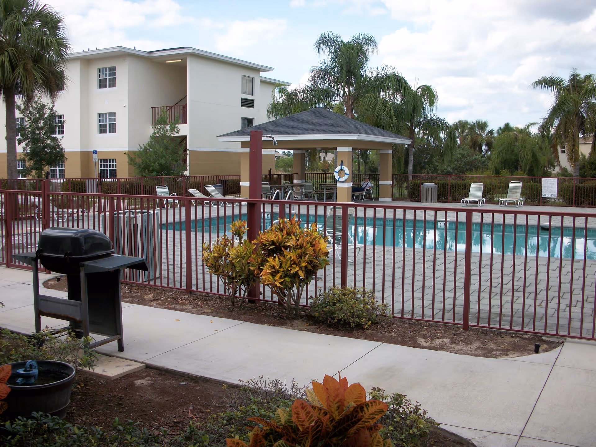 Outdoor swimming pool area at Heron Pond Apartments with a fenced pool, lounge chairs, a covered seating area, palm trees, and a barbecue grill on a concrete pathway.