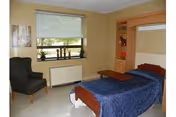 Small nursing home bedroom featuring a single bed with blue cover, chair, window, and storage unit.