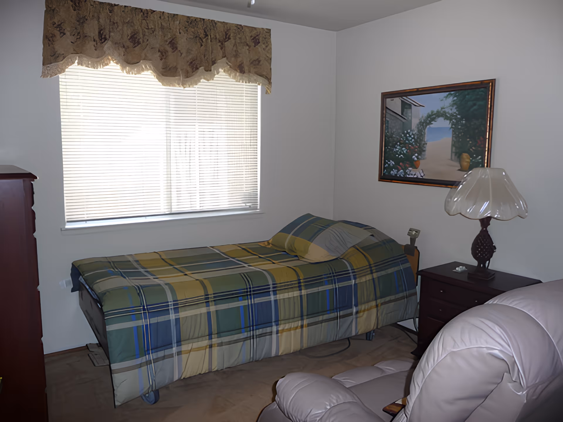 A small bedroom with a single bed covered in a green, blue, and yellow plaid bedspread. There is a window with blinds and a decorative valance above it. Next to the bed is a dark wooden nightstand with a lamp that has a white, translucent lampshade. A framed painting of a garden archway is hanging on the wall above the nightstand. In the foreground, part of a light-colored recliner chair is visible.