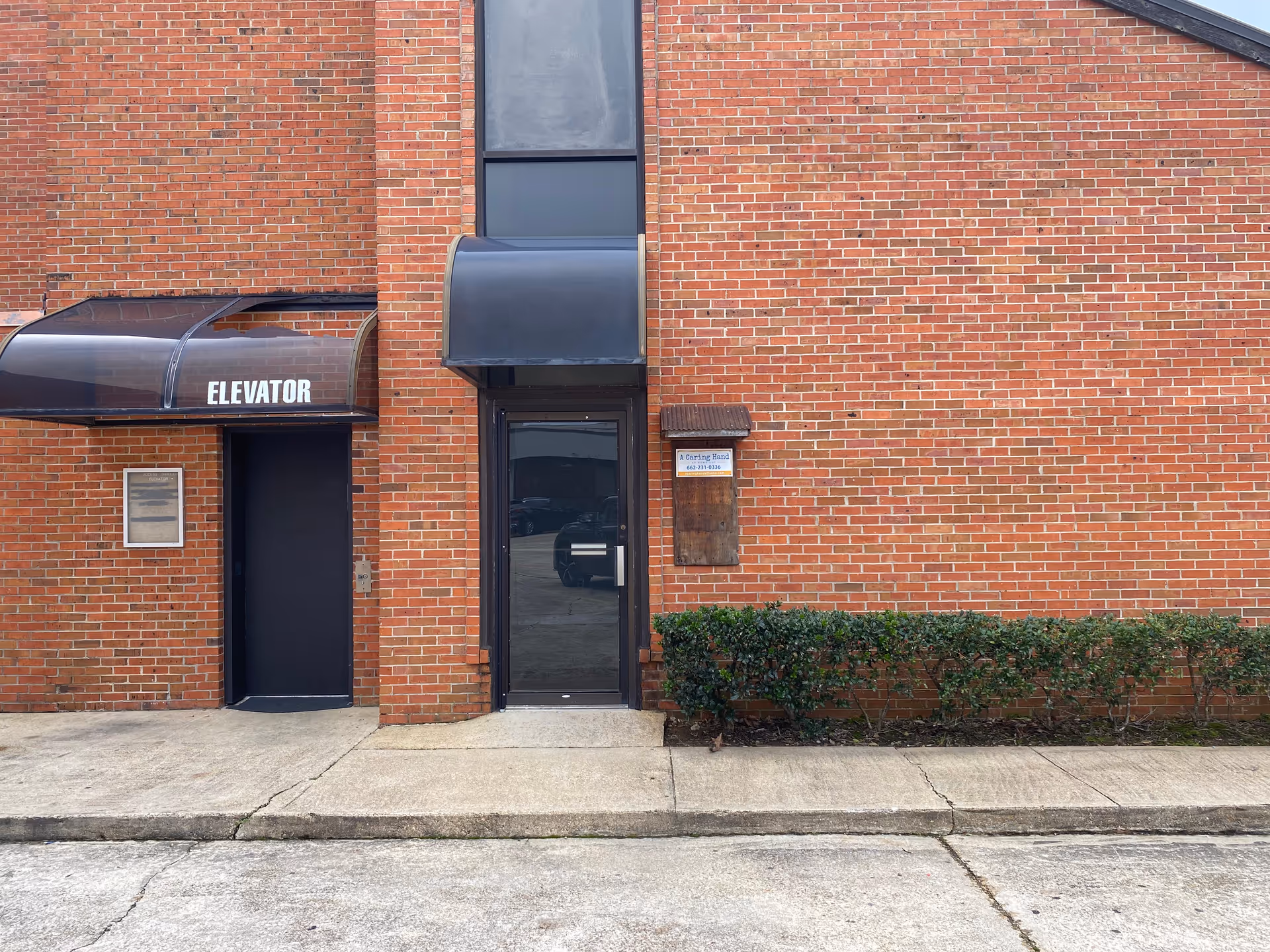 Brick facility exterior with entrance doors, signage, and shrub-lined sidewalk.
