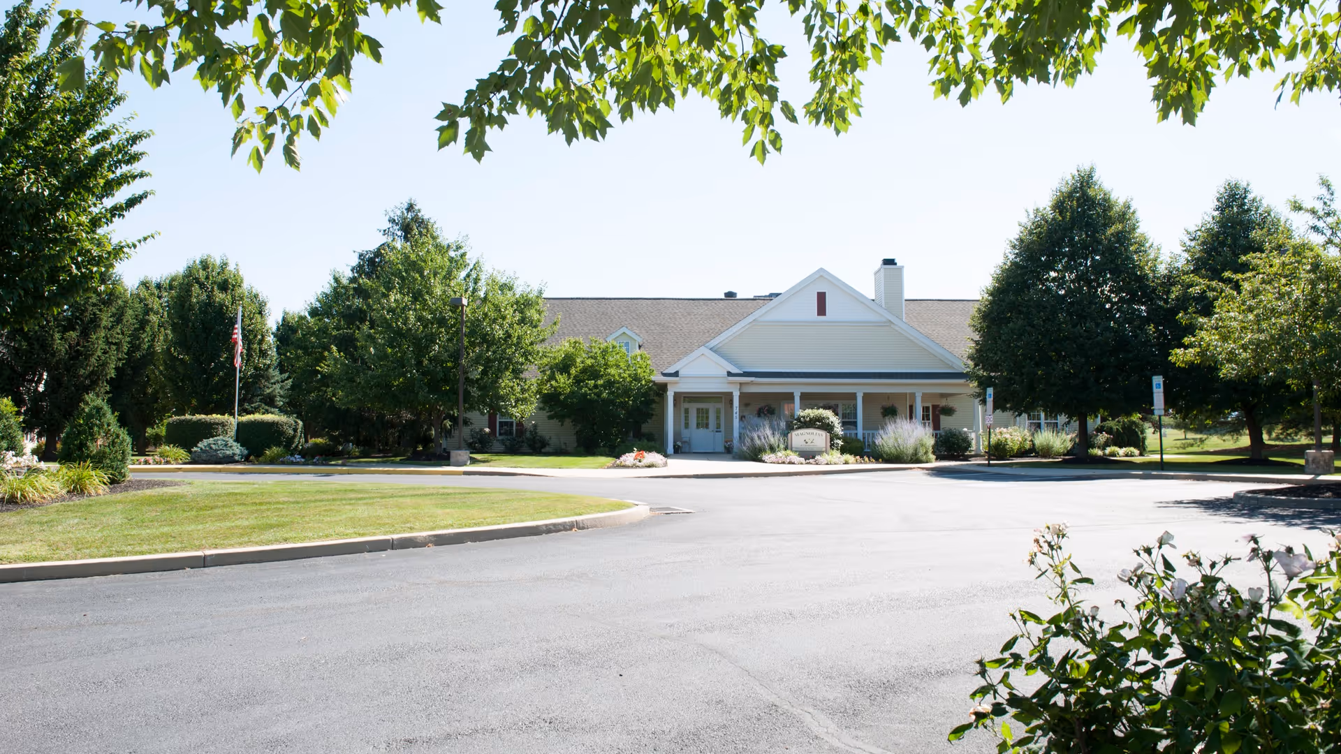 Front exterior view of a single-story building with a covered entrance, surrounded by well-maintained landscaping including trees, bushes, and a lawn under a clear sky.