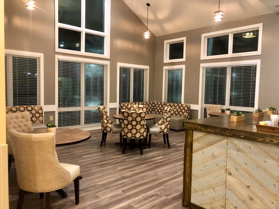 A cozy interior common area with multiple seating options including patterned chairs around a round table, a beige armchair, and a corner bench with circular patterned upholstery. The room has large windows with white blinds, high ceilings with pendant lights, and wood-look flooring. A wooden counter with small potted plants is visible on the right side.