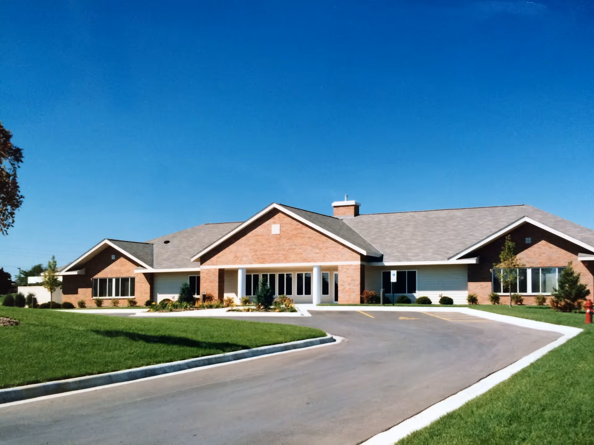 Exterior front view of a single-story brick building with a gray shingled roof, white columns at the entrance, large windows, a driveway, and well-maintained green lawn under a clear blue sky.