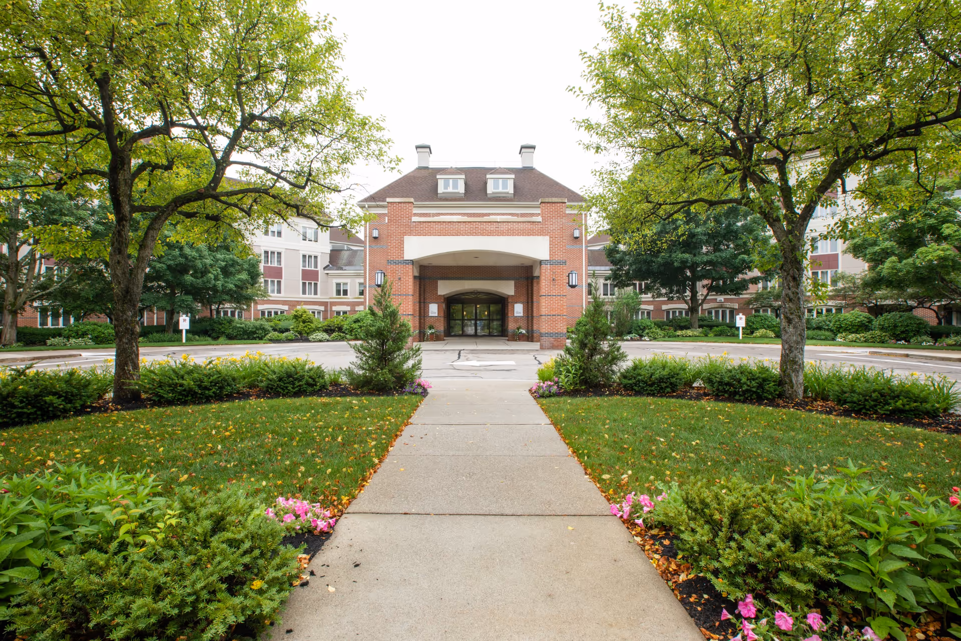 Brick senior living facility with central entrance, walkway, and lush landscaping.
