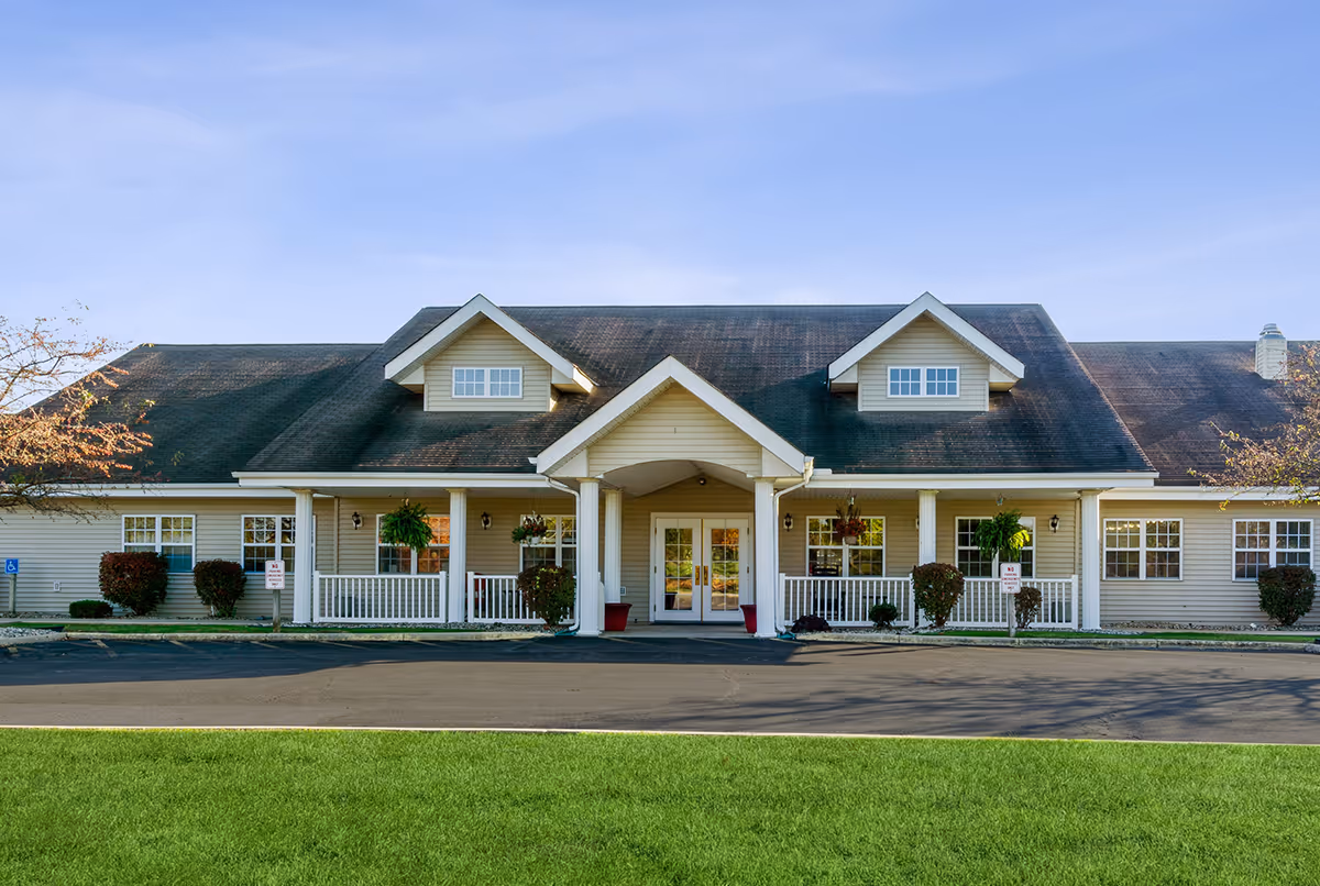 Front exterior view of Cedar Creek of Marion facility, showing a single-story building with beige siding, a dark roof, white columns supporting a covered entrance, hanging plants, and neatly trimmed bushes. The sky is clear and blue, and there is a paved driveway and green lawn in front.