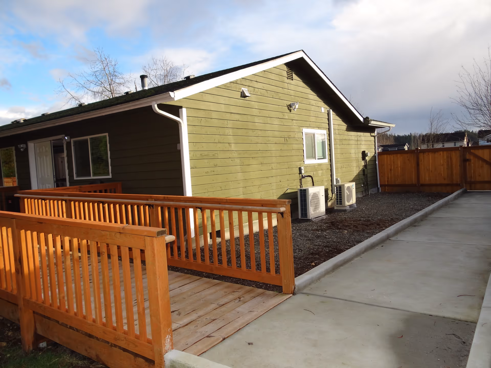 Green single-story building exterior with wooden deck, ramp, brown fence, and AC units.