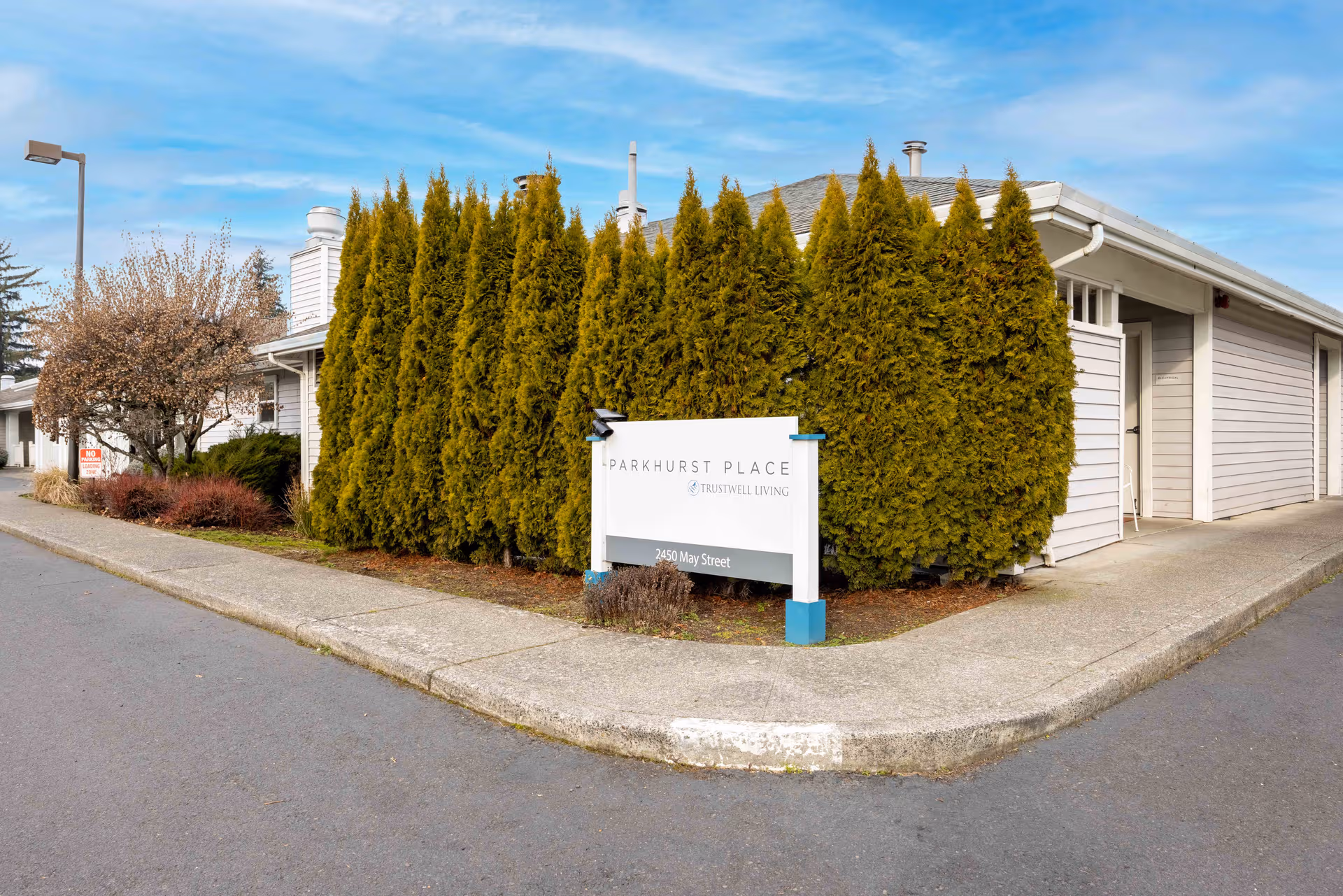 Front exterior of Trustwell Living at Parkhurst Place with tall hedges and a sign by curb.