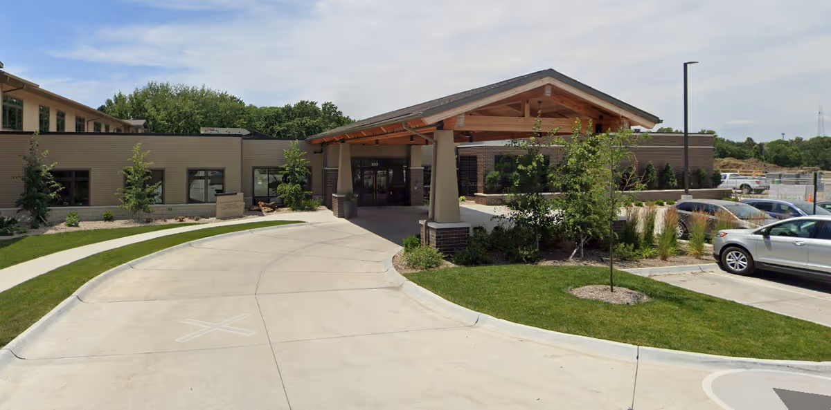 Front entrance of a senior living facility with a covered porte-cochere, driveway, landscaped beds, and parked cars.