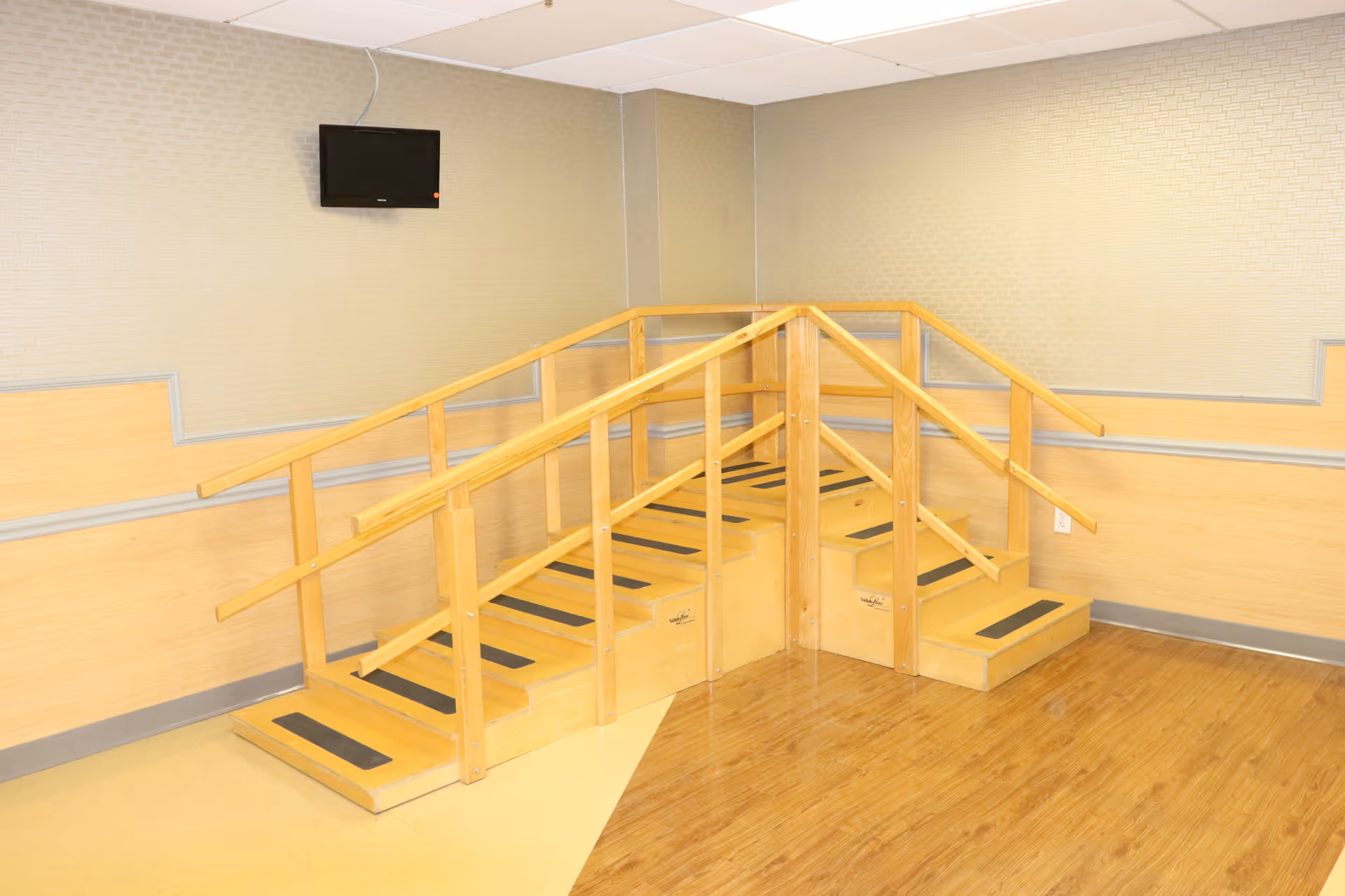 Indoor senior living area featuring a wooden staircase with handrails and a wall-mounted TV.
