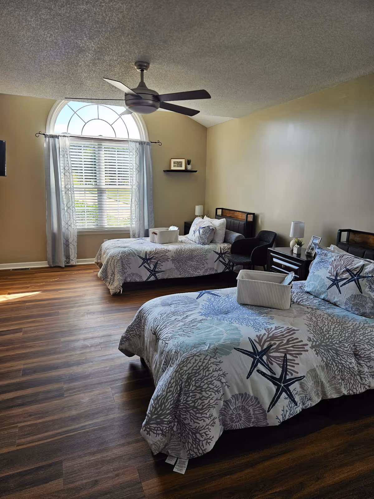Two twin beds with ocean-themed bedding and a large arched window in a wood-floored bedroom.