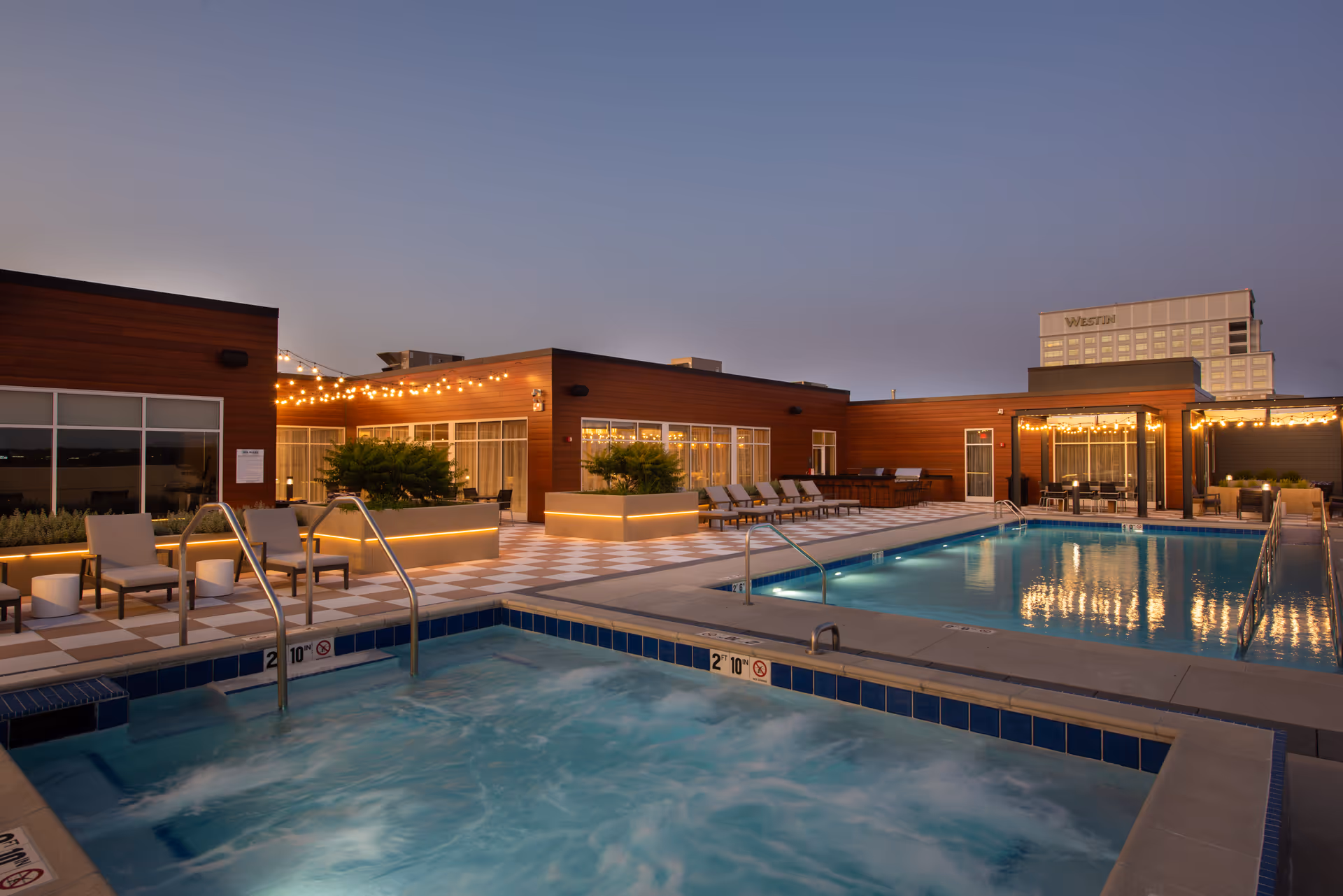 Rooftop pool and hot tub area at dusk with lounge chairs, string lights, and a modern building facade.