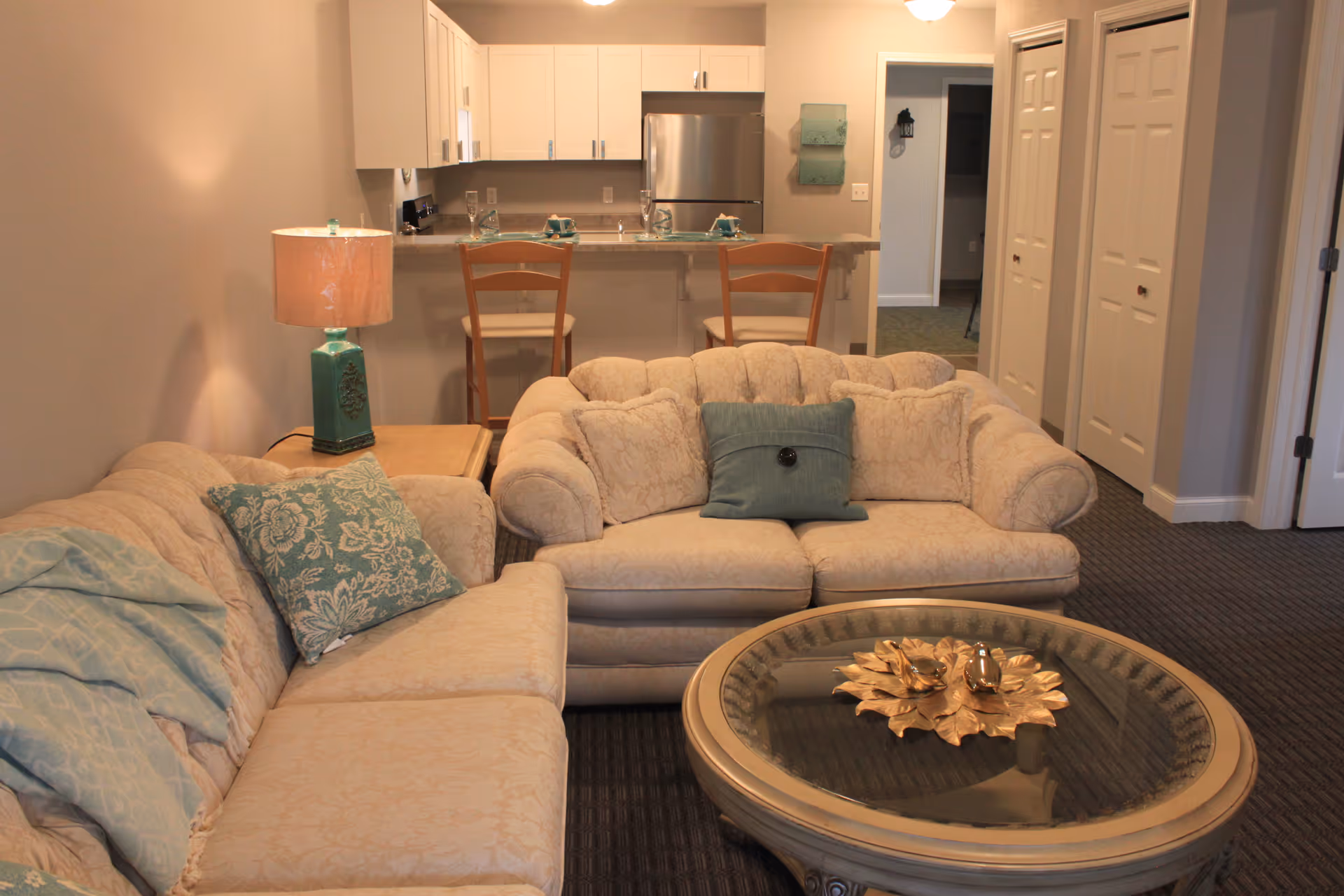 A cozy living room area with two beige upholstered sofas adorned with blue and patterned cushions. A round glass coffee table with a decorative centerpiece sits in front of the sofas. Behind the seating area is a kitchen counter with two wooden chairs, white cabinets, and a stainless steel refrigerator. A turquoise lamp is on a side table next to one of the sofas. The room has neutral-colored walls and carpeted flooring.