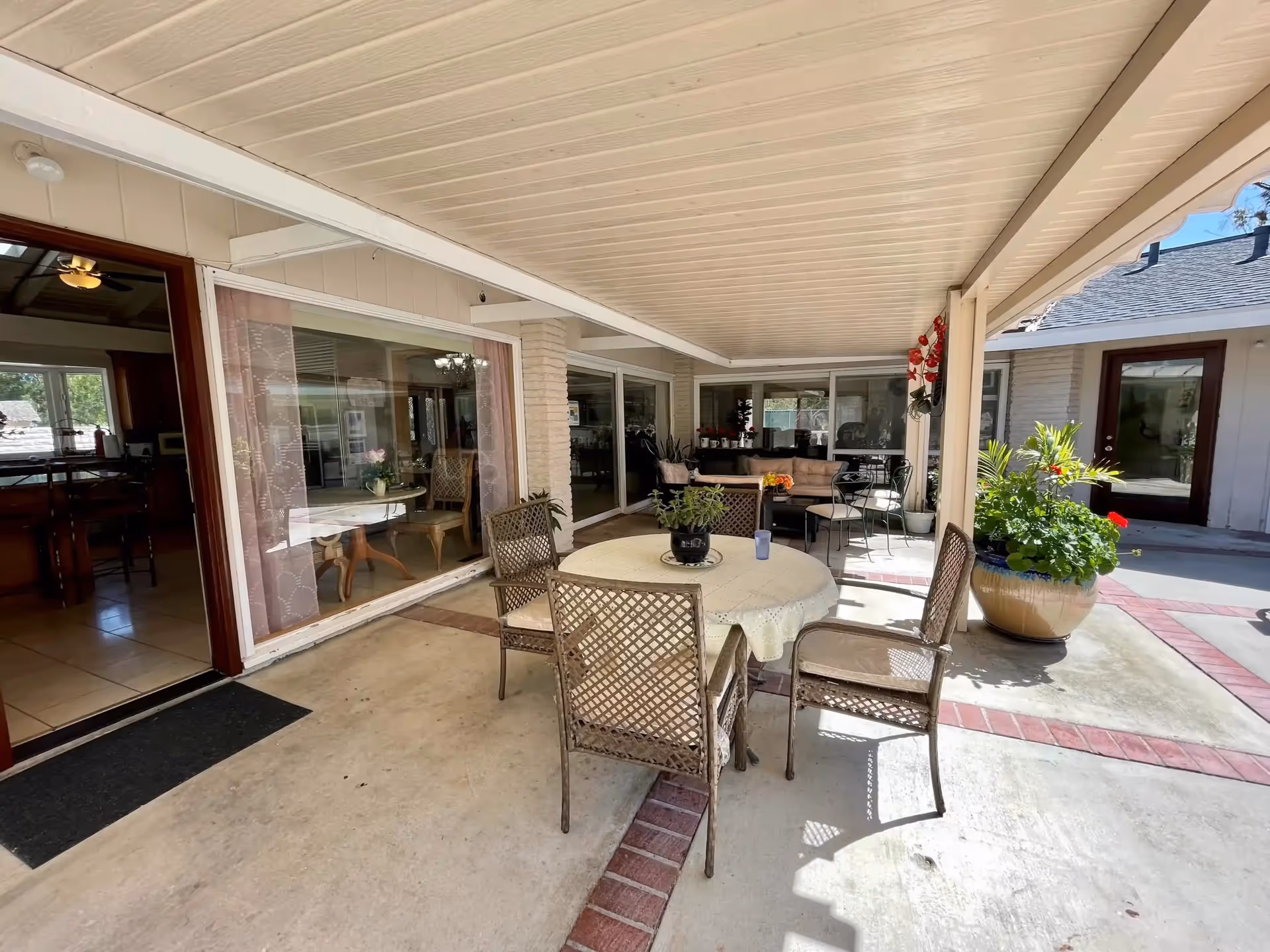 Covered outdoor patio with round table, chairs, potted plants, and glass doors to indoor rooms.