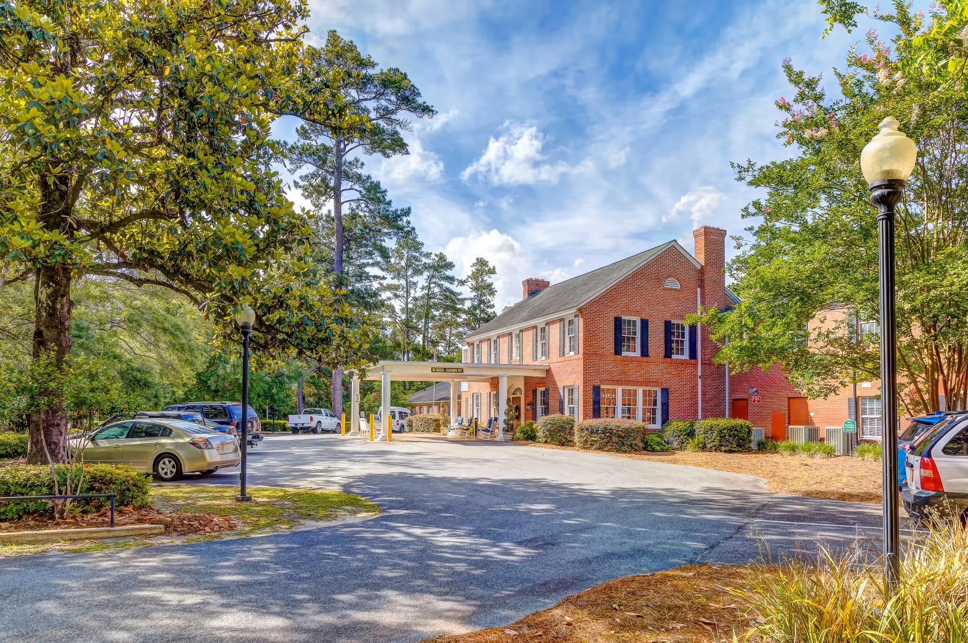 Brick two-story senior living building with a covered entrance, parking lot, trees, and lamp posts under a blue sky.