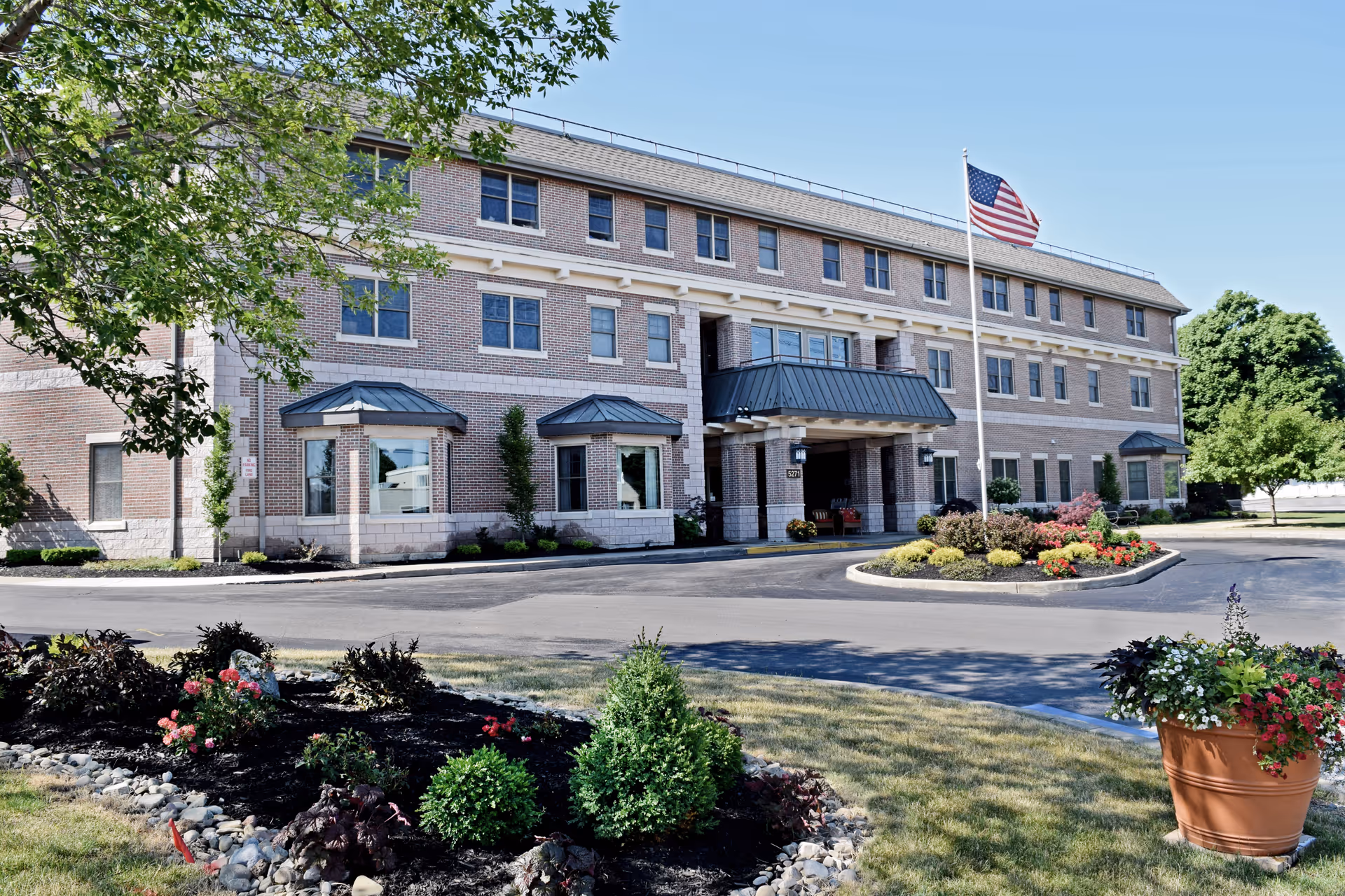 Exterior view of a three-story brick senior living facility building with a covered entrance, an American flag on a flagpole, landscaped flower beds, and trees under a clear blue sky.