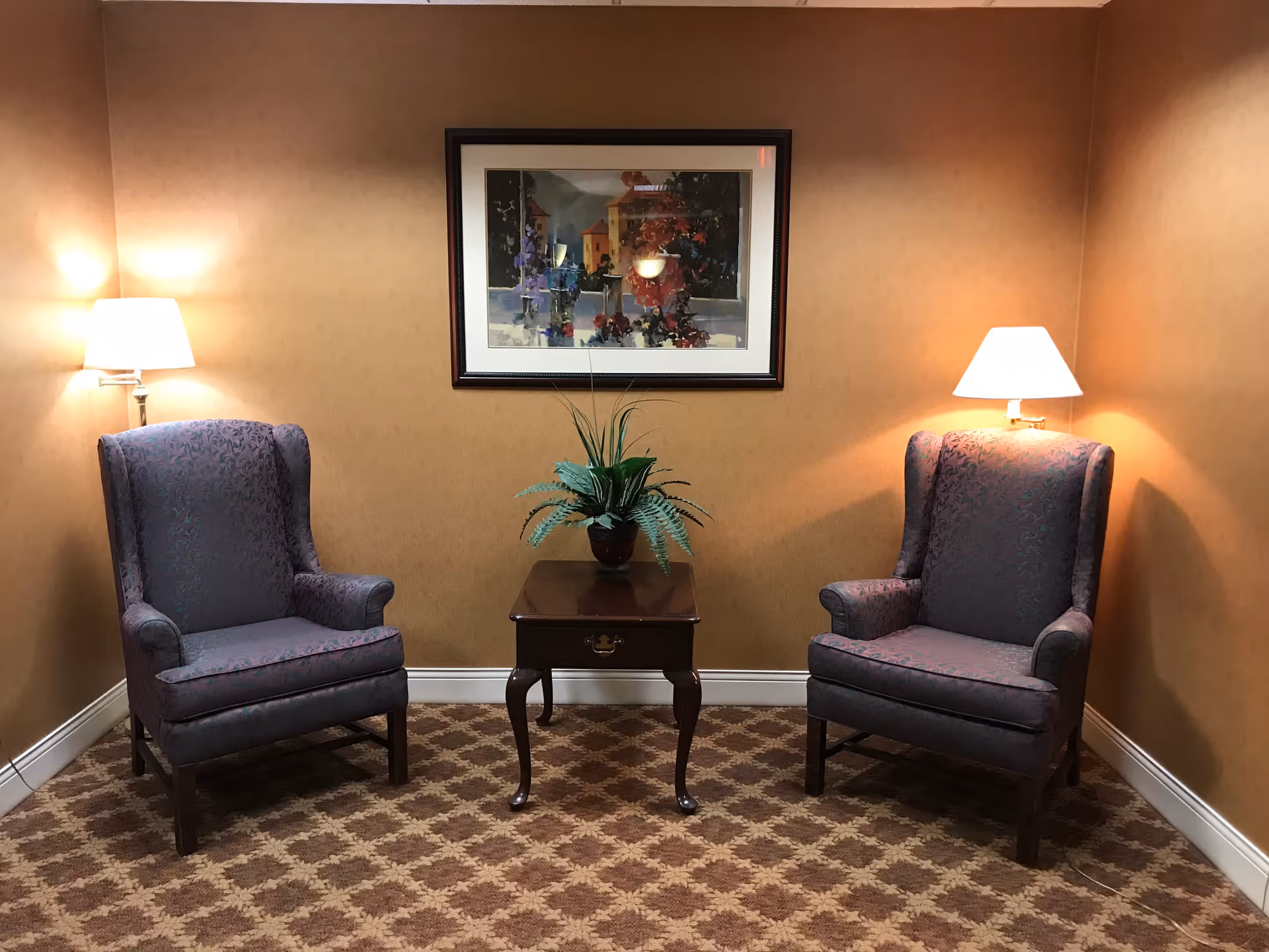 A cozy corner with two upholstered armchairs in a patterned fabric, positioned on either side of a small wooden side table with a potted plant. The walls are painted a warm beige color, and there are two wall-mounted lamps above each chair providing soft lighting. A framed painting hangs on the wall above the table.