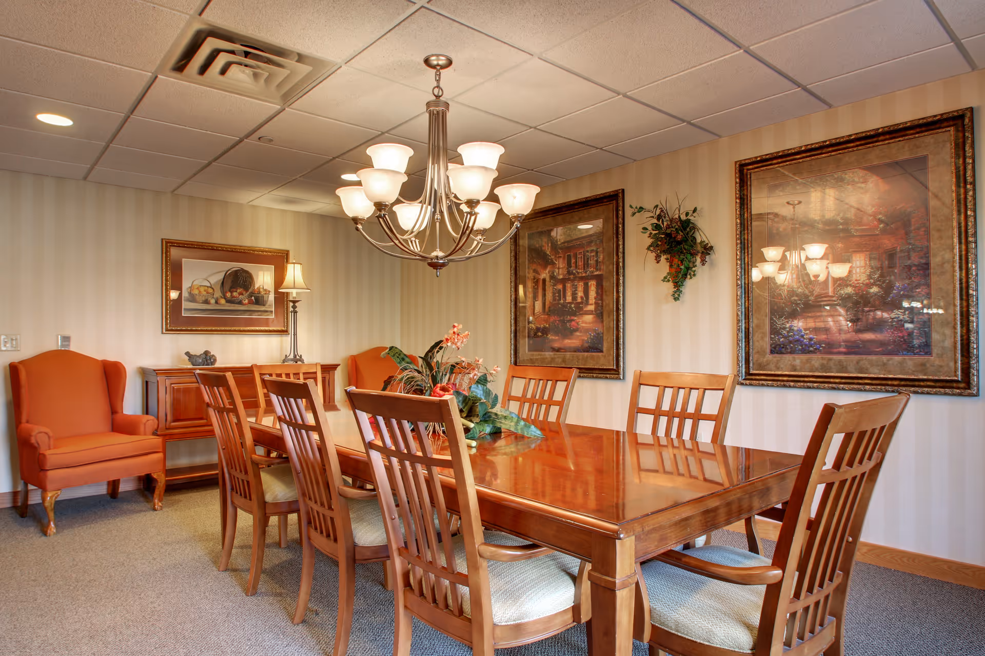 A dining room with a large wooden table surrounded by eight wooden chairs with cushioned seats. The room features a chandelier with multiple lights hanging above the table, two framed paintings on the wall, a small sideboard with a lamp and decorative items, and an orange upholstered armchair in the corner. The walls have a light striped wallpaper and there is a small plant hanging on the wall.