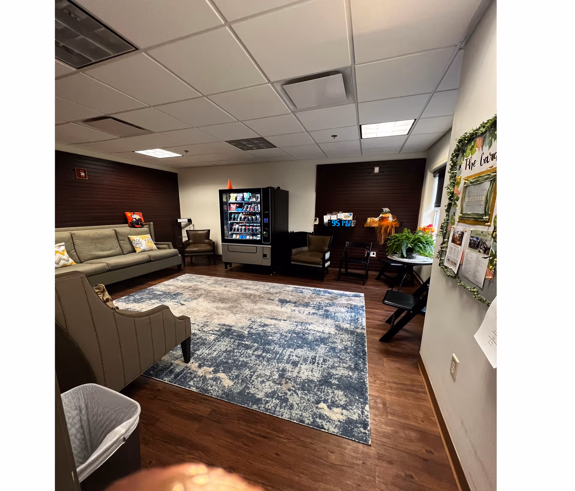 Indoor senior living lounge with couches, chairs, a vending machine, and a blue rug.