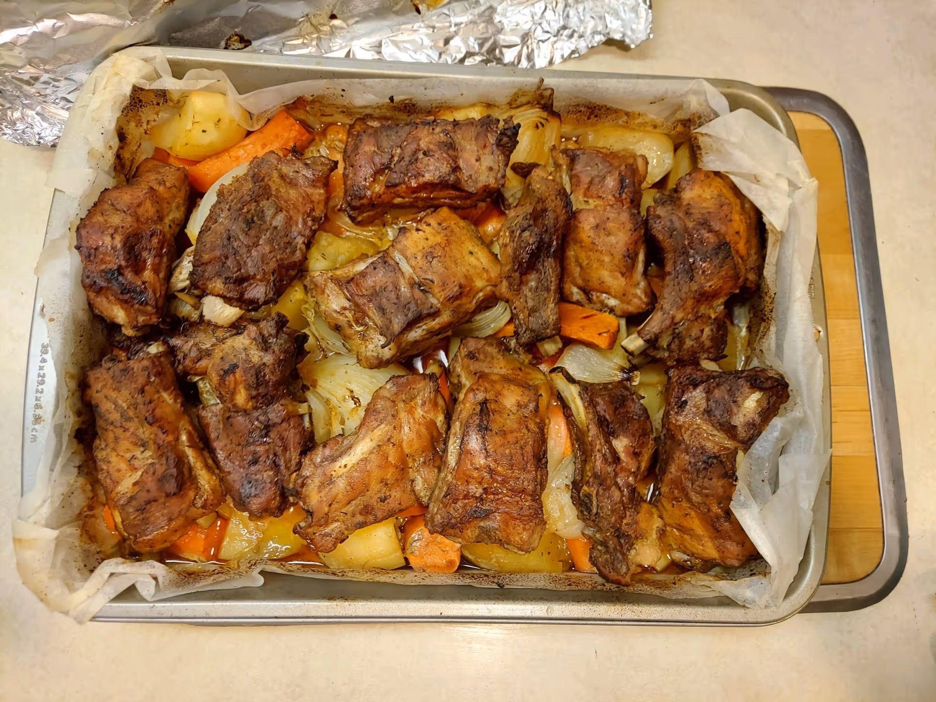 A baking tray lined with parchment paper containing roasted pork ribs with cooked vegetables including carrots, onions, and potatoes.
