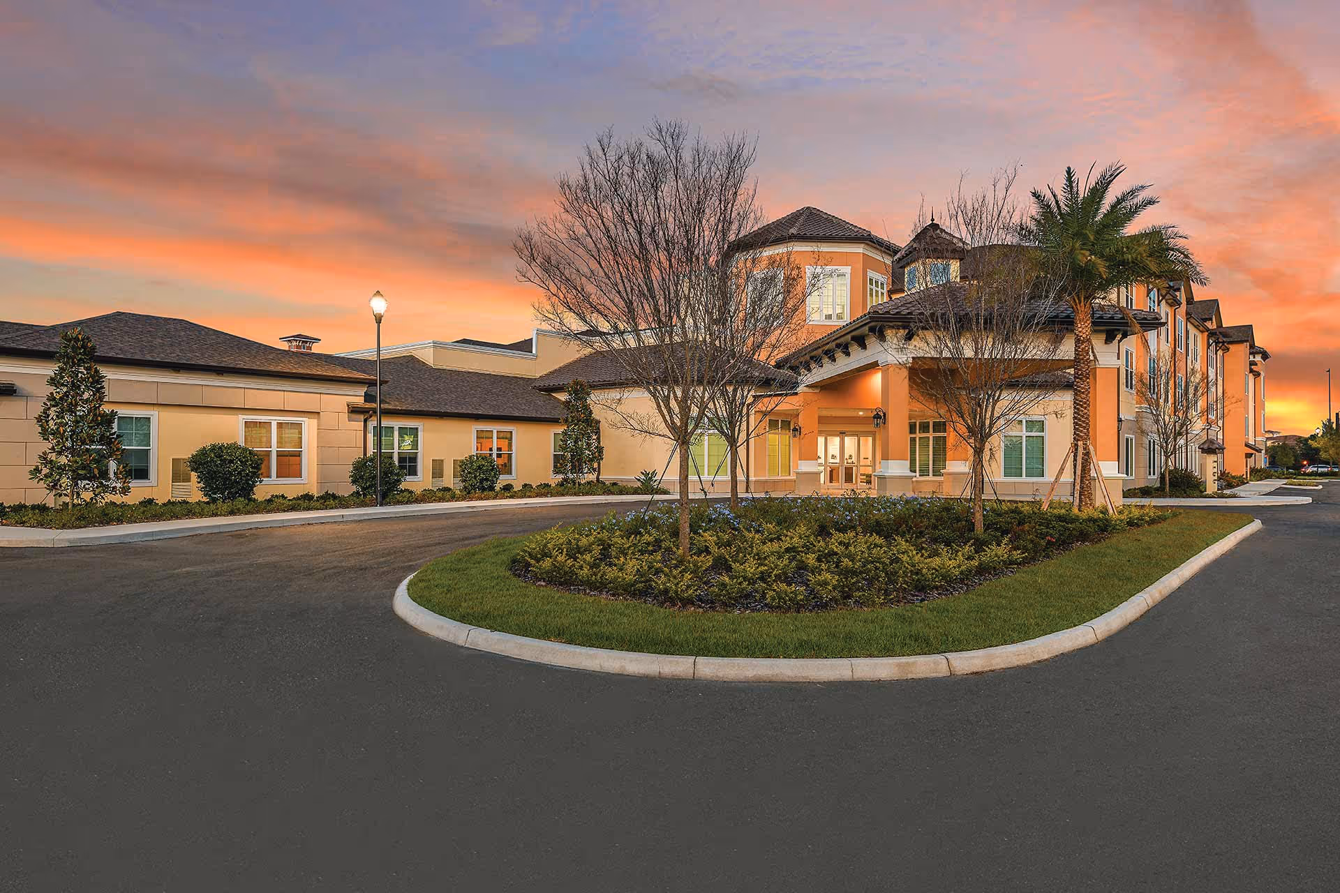 Front exterior of a multi-story senior living building with a landscaped circular driveway under a colorful sunset sky.