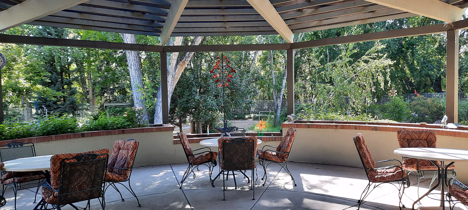 Outdoor covered patio area with several round tables and cushioned metal chairs. The patio overlooks a garden with trees and greenery, providing a shaded and relaxing environment.