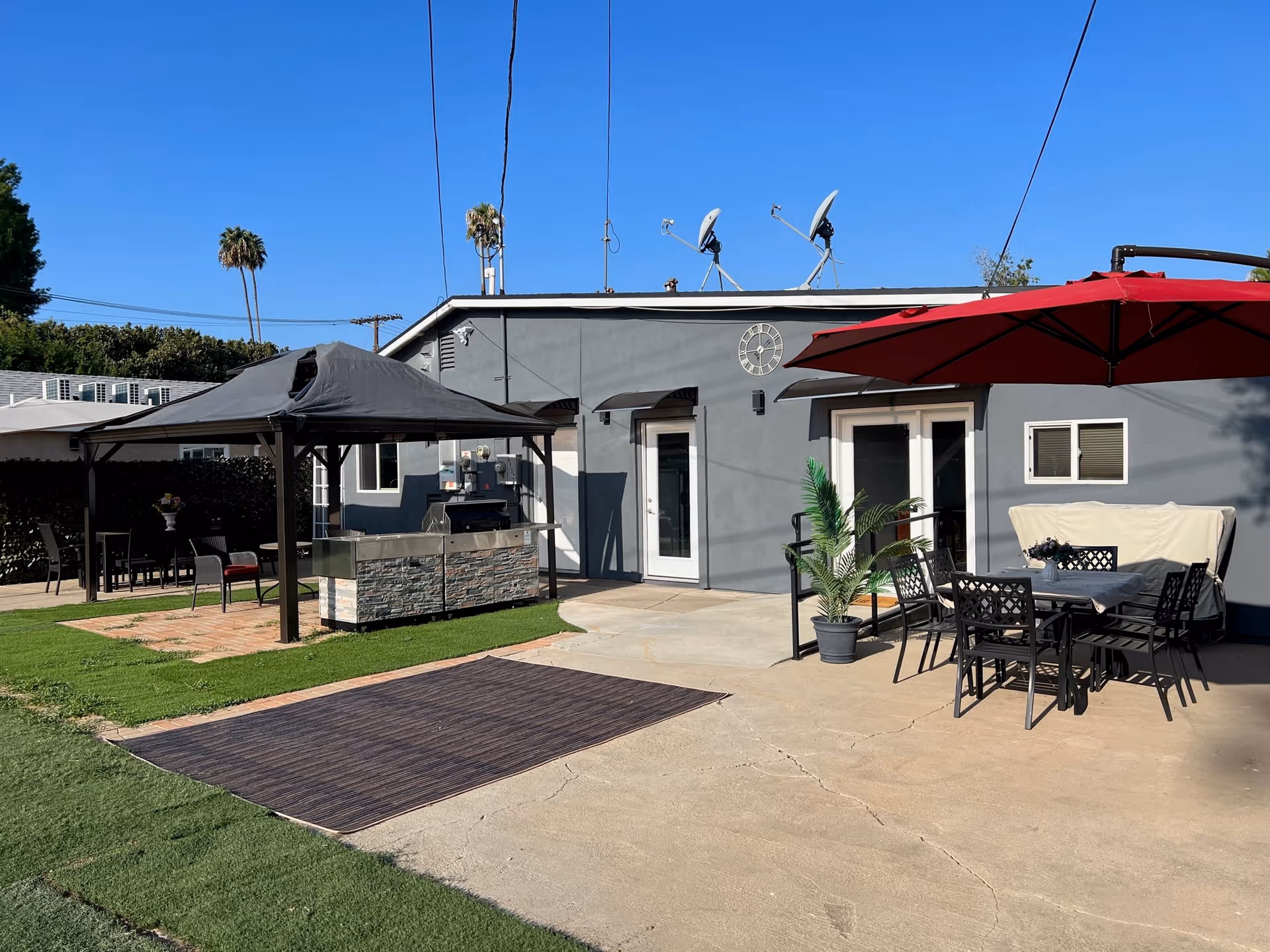 Outdoor patio area with seating, umbrellas, a covered grill station, and a gray building façade under a clear blue sky.