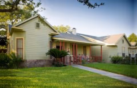 Exterior view of a single-story residential building with light green siding, a covered porch with red pillars, and a well-maintained lawn with trees in the background under a clear sky.