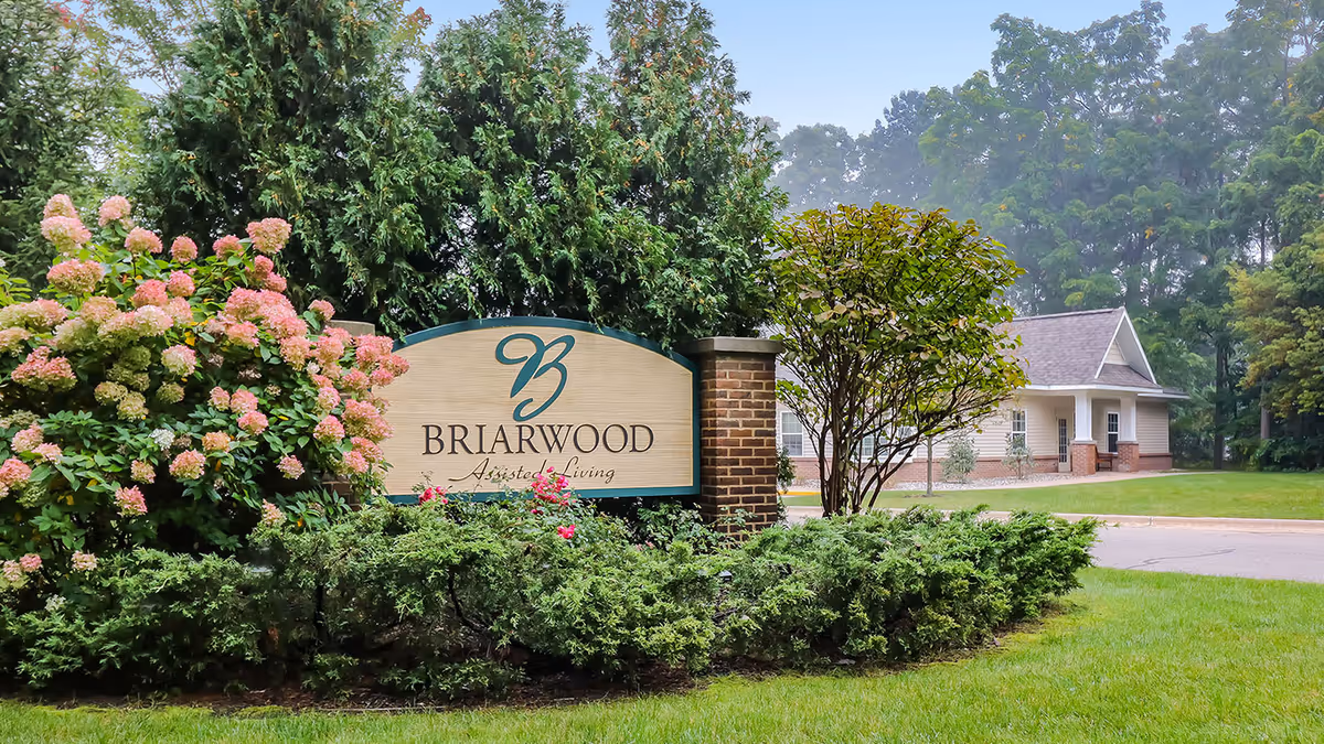 A Briarwood Assisted Living entrance sign surrounded by flowering shrubs and greenery with the facility building and trees in the background.