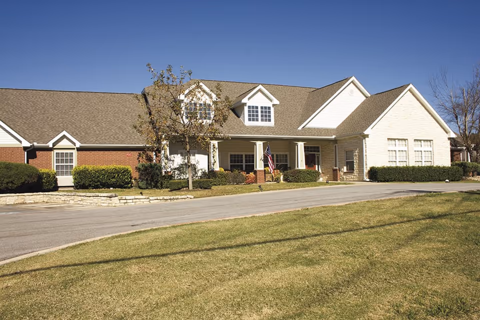 Single-story brick and stone senior living building with gabled roofs, a front porch displaying an American flag, driveway, and grassy lawn under a clear blue sky.