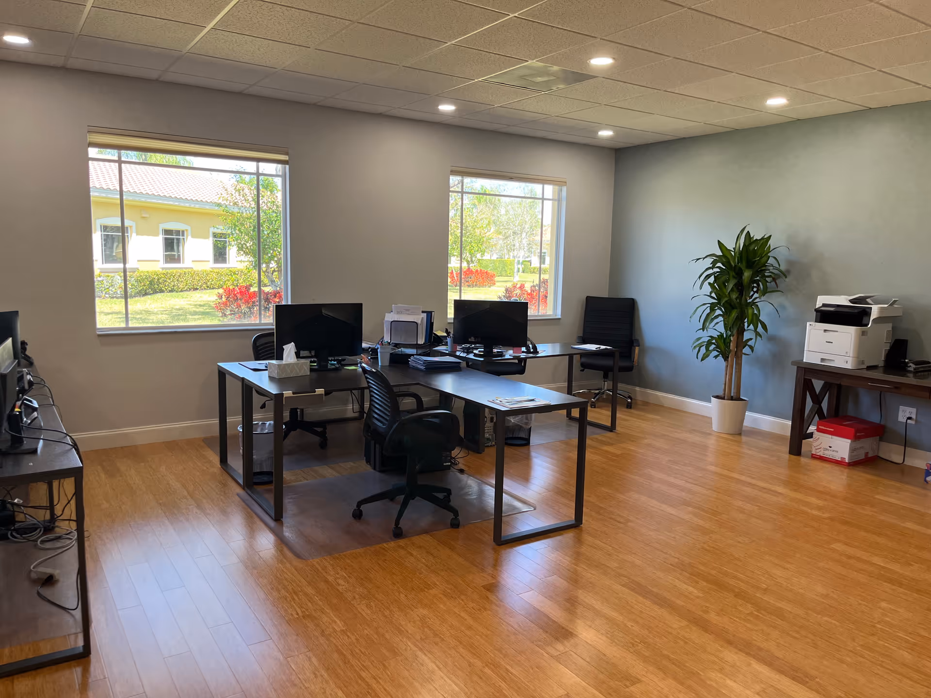 Office workspace with desks, computers, printer, and a plant in a bright, modern room.