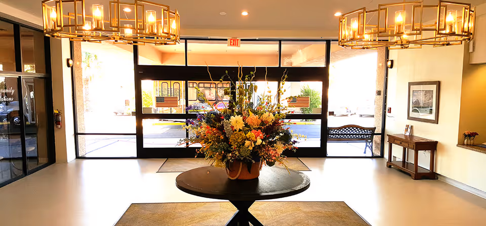 Entrance lobby of a senior living facility with a round table in the center holding a large floral arrangement. The lobby has glass doors leading outside, two modern chandeliers hanging from the ceiling, a bench near the door, and a small wooden table with a framed picture on the wall.