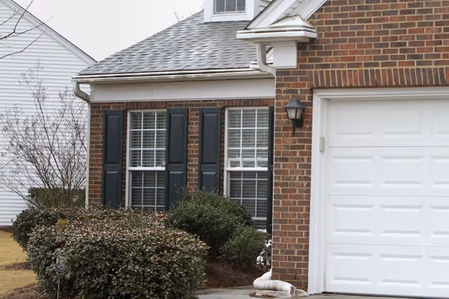 Brick residential home exterior with window shutters, shrubs, and a white garage door.