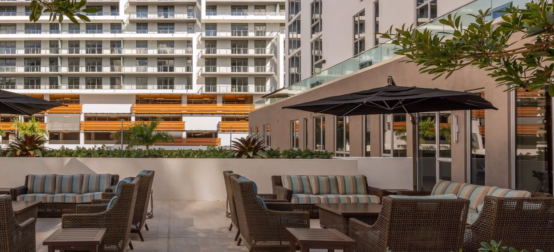 Outdoor courtyard with wicker lounge chairs and sofas with striped cushions, patio umbrellas, and a multi-story residential building in the background.