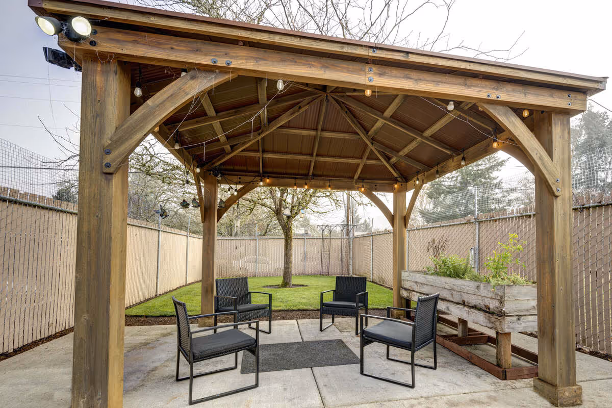 Outdoor seating area under a wooden gazebo with four black chairs arranged around a small black rug. The area is surrounded by a tall fence with netting, and there is a tree and a raised planter box with plants in the background.