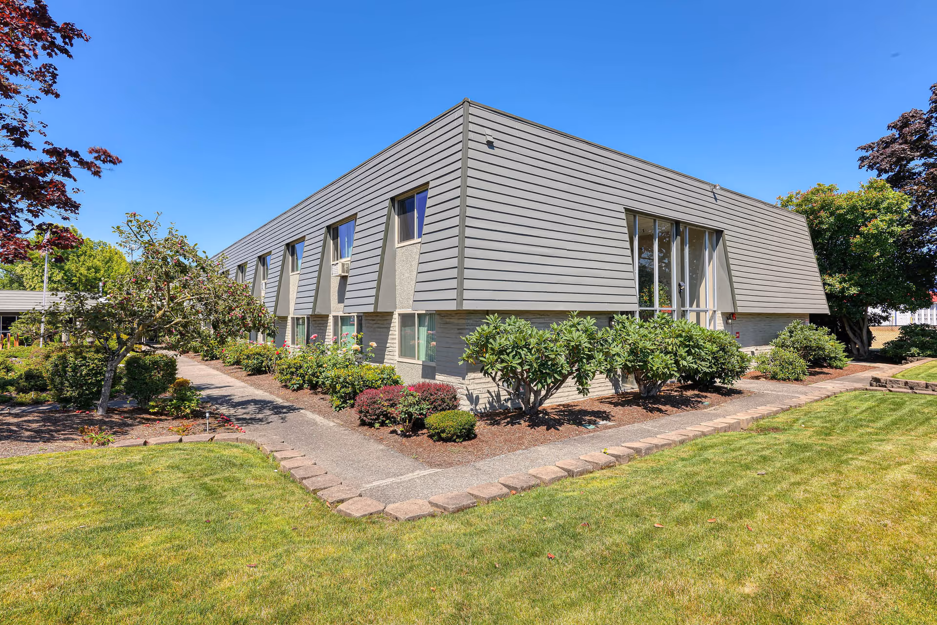 Gray two-story retirement facility building with landscaped shrubs, a walkway, and lawn beneath a clear blue sky.