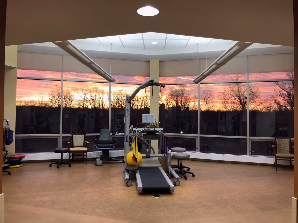 Interior gym room with treadmill and equipment, sunset-lit large windows.