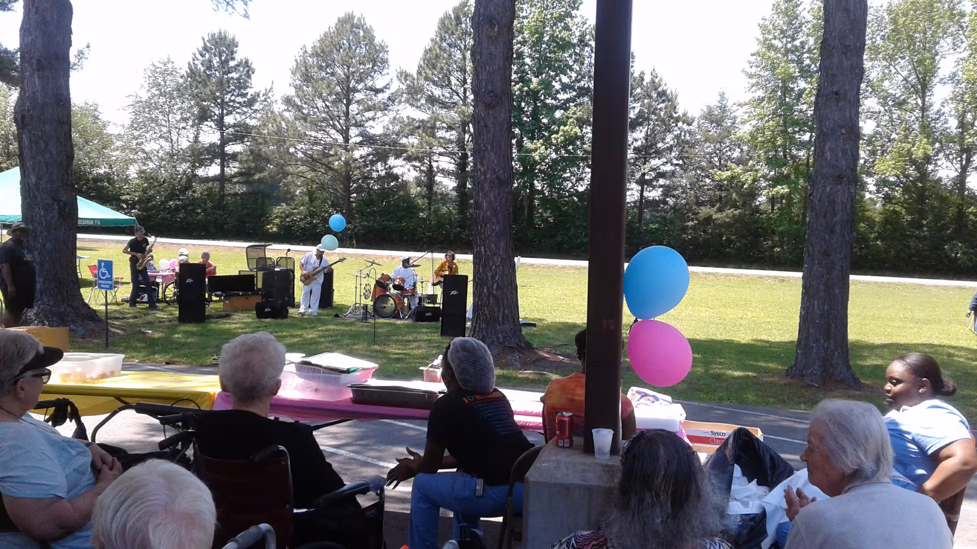 Residents watch a live band outdoors under trees at an outdoor park-style event.