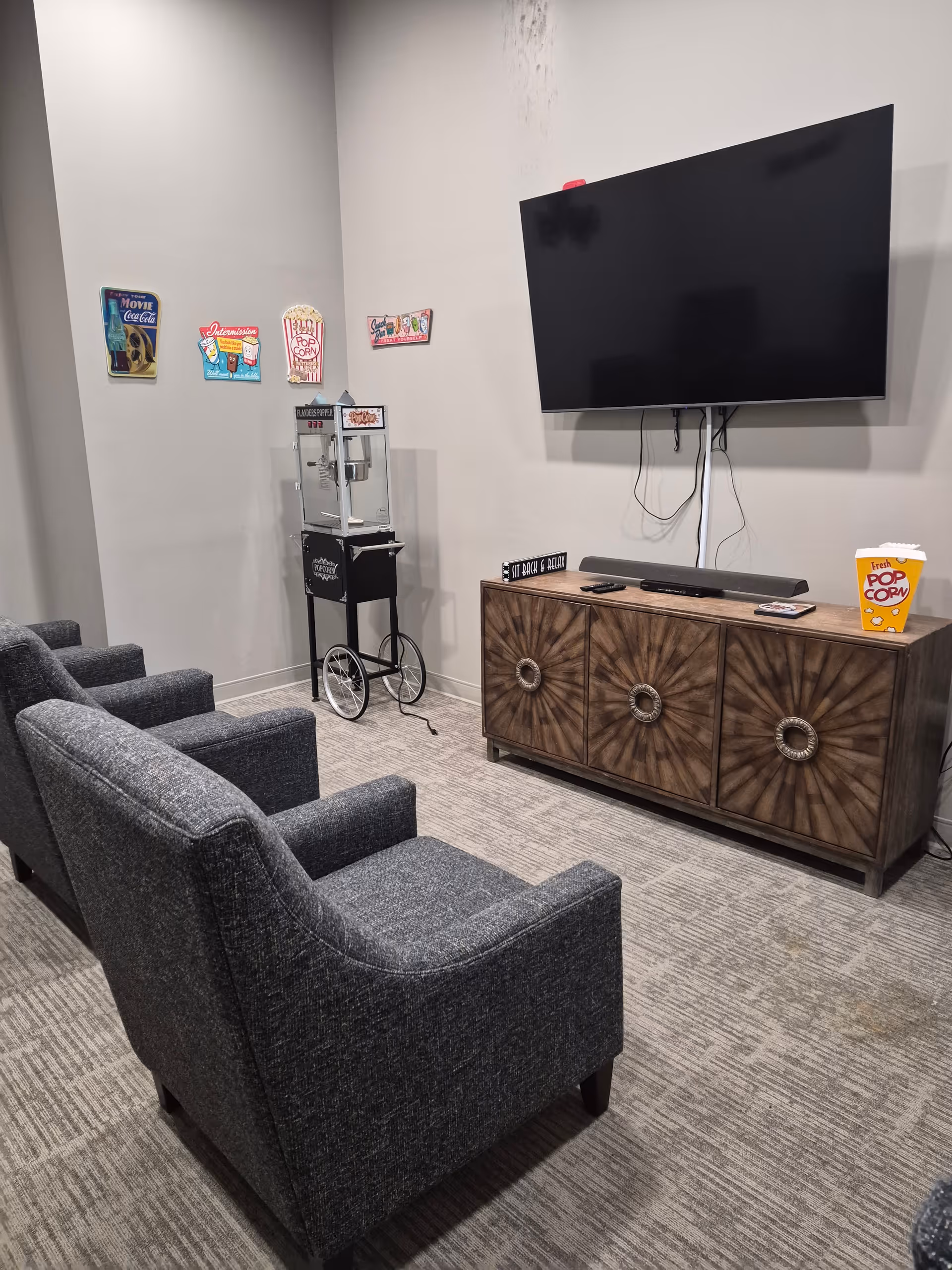 Cozy lounge area with gray chairs, a mounted TV, posters, and a popcorn machine.