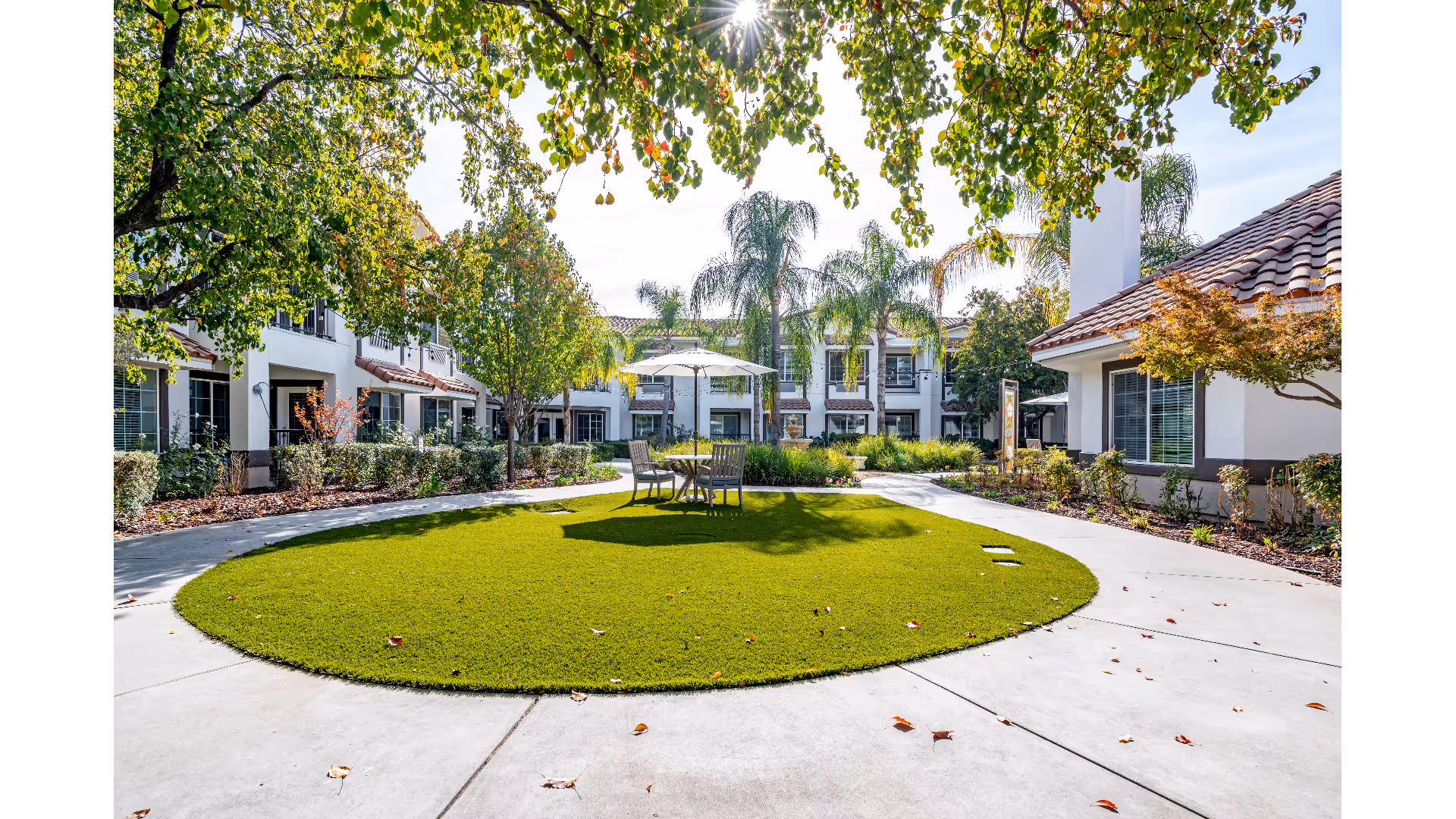 Sunny central courtyard with a circular lawn, a table and umbrella, surrounded by two-story Mediterranean-style buildings and palm trees.