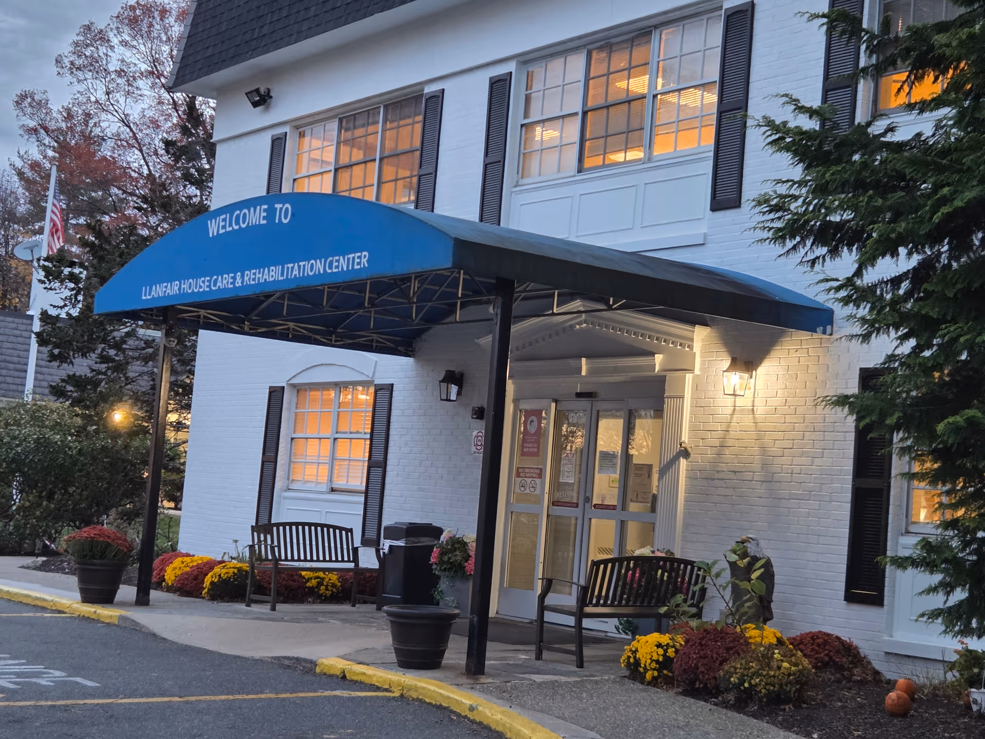 White brick care facility with blue awning entrance and benches, colorful flowers outside.