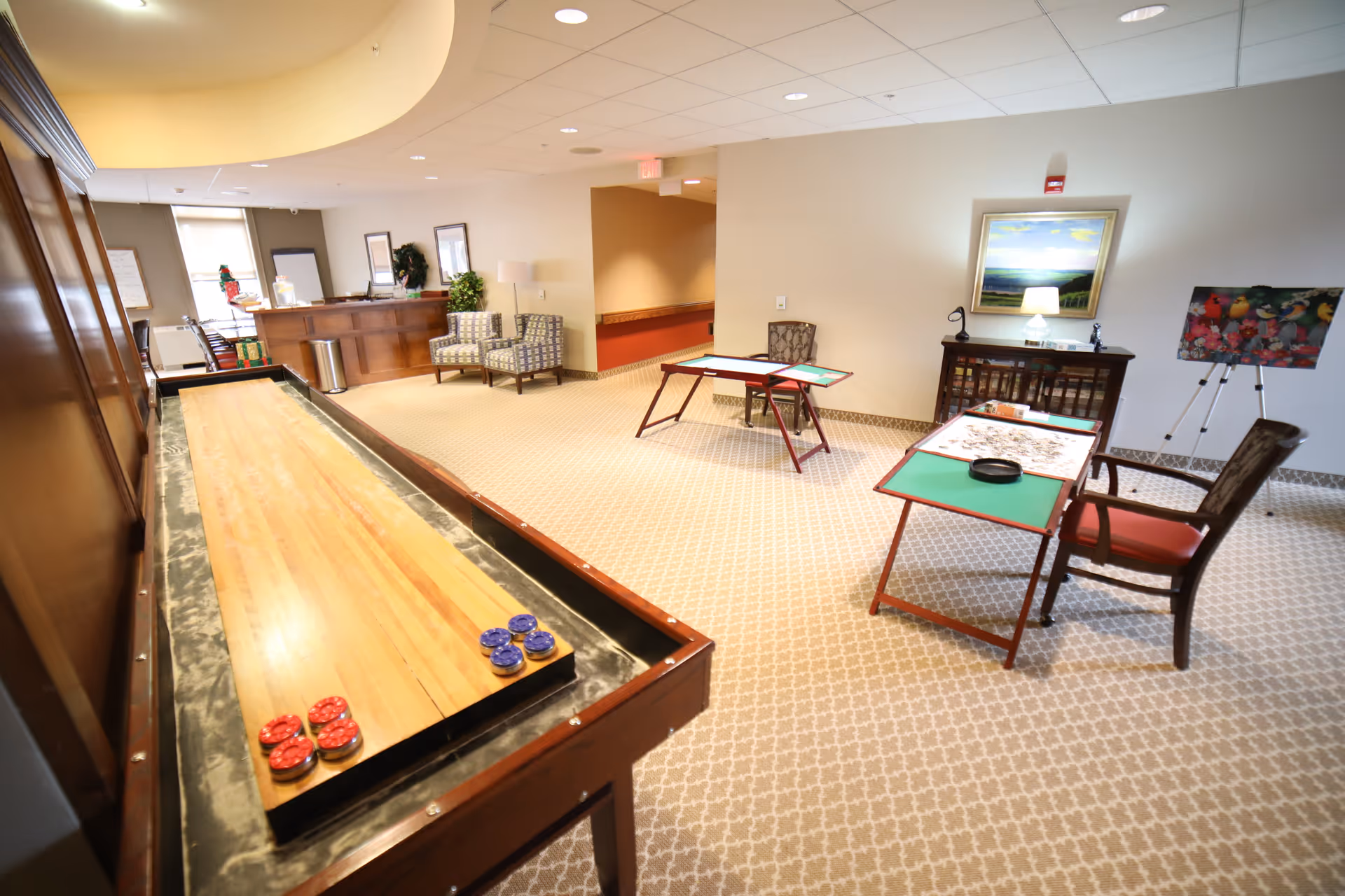 Bright communal activity room in a senior living facility featuring a shuffleboard table, game tables, chairs, and a reception desk.