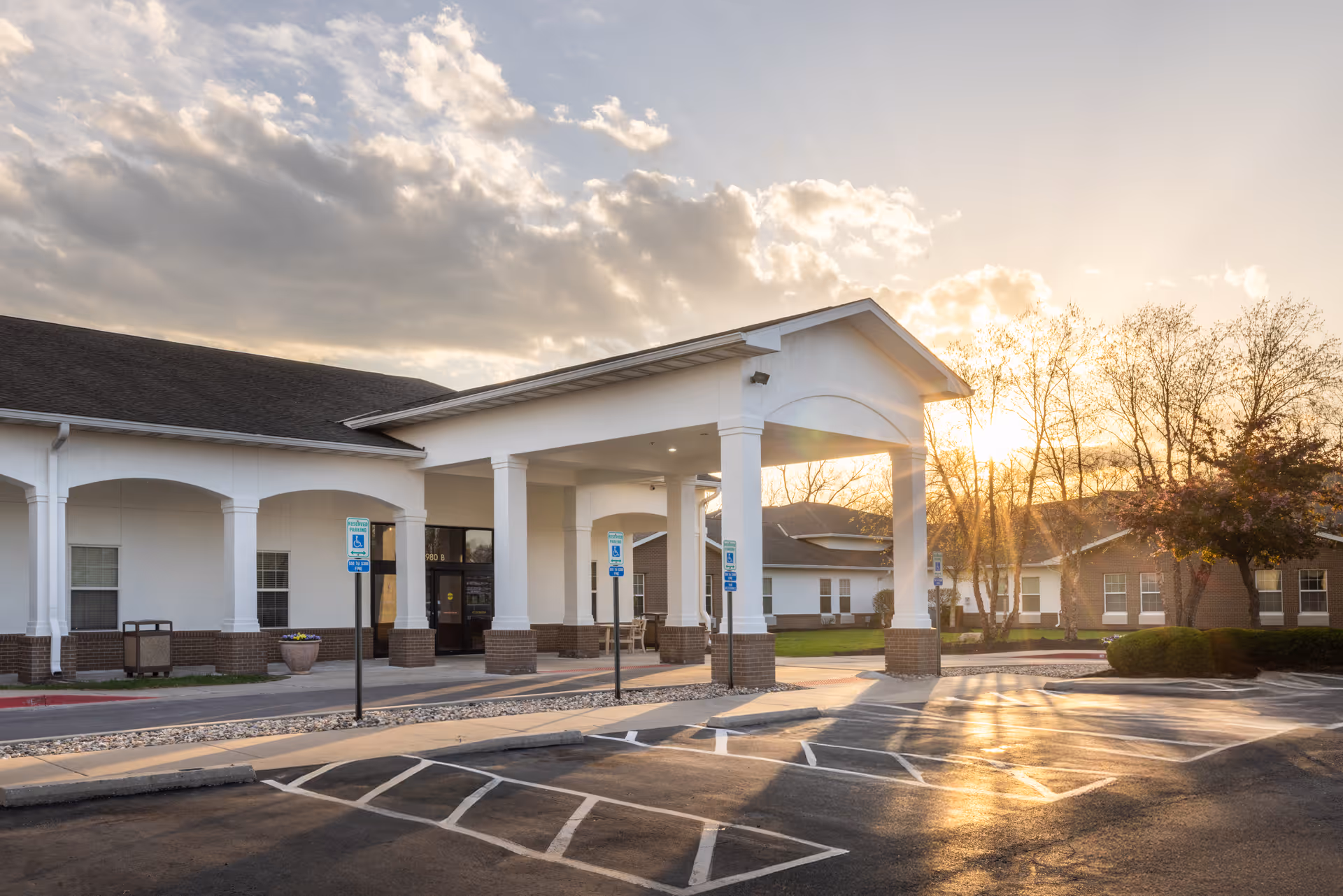Exterior view of Jackson Creek Post Acute facility at sunset, showing the entrance with a covered drop-off area, handicap parking spaces, and surrounding trees with a partly cloudy sky.