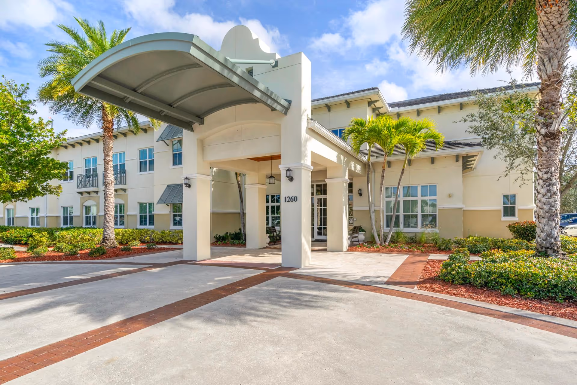 Front entrance of a two-story senior living building with a covered porte-cochere, palm trees, and landscaped grounds.