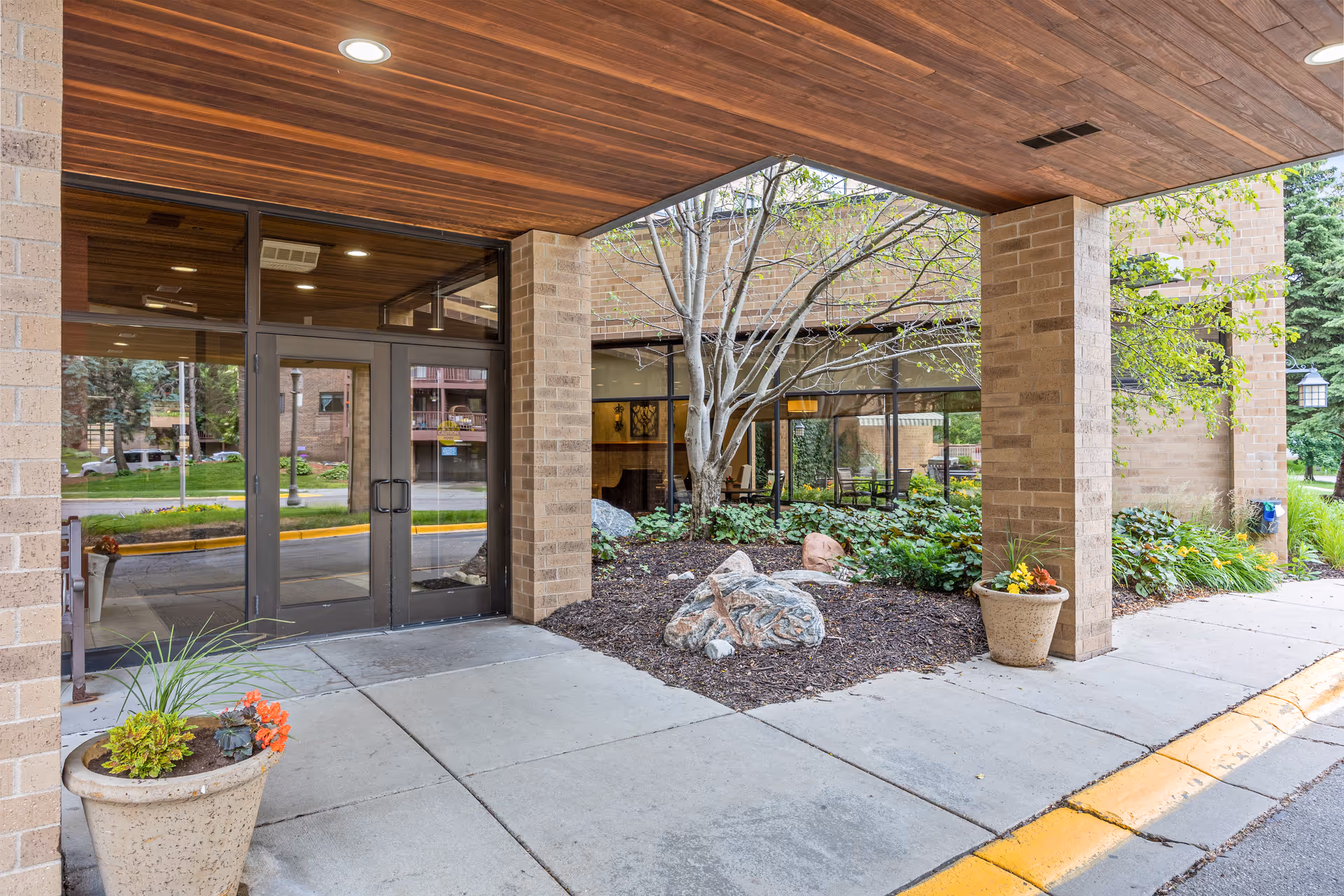 Entrance area of The Wellington Senior Living facility showing glass double doors, brick pillars, a covered walkway with a wooden ceiling, potted plants, and landscaping with rocks and greenery.