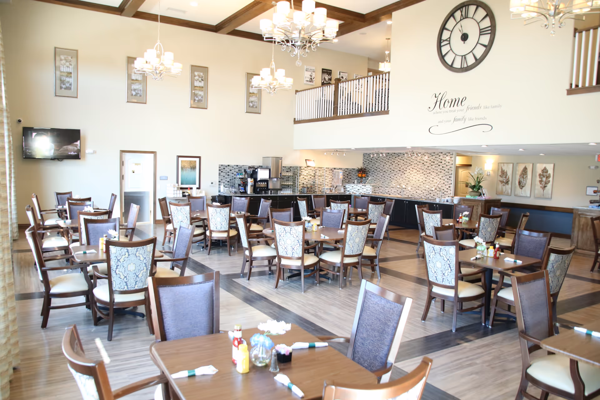 Spacious, well-lit dining room with numerous wooden tables and upholstered chairs, chandeliers, and a buffet counter under a high ceiling.
