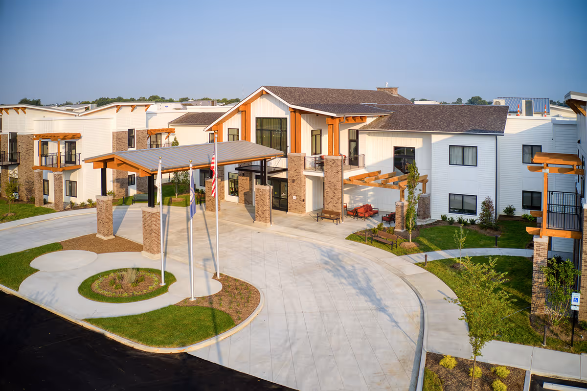 Exterior view of Preston Greens Senior Living facility showing a modern two-story building with white siding, brick accents, and wooden beams. The entrance features a covered drop-off area with three flagpoles and landscaped greenery around the driveway.