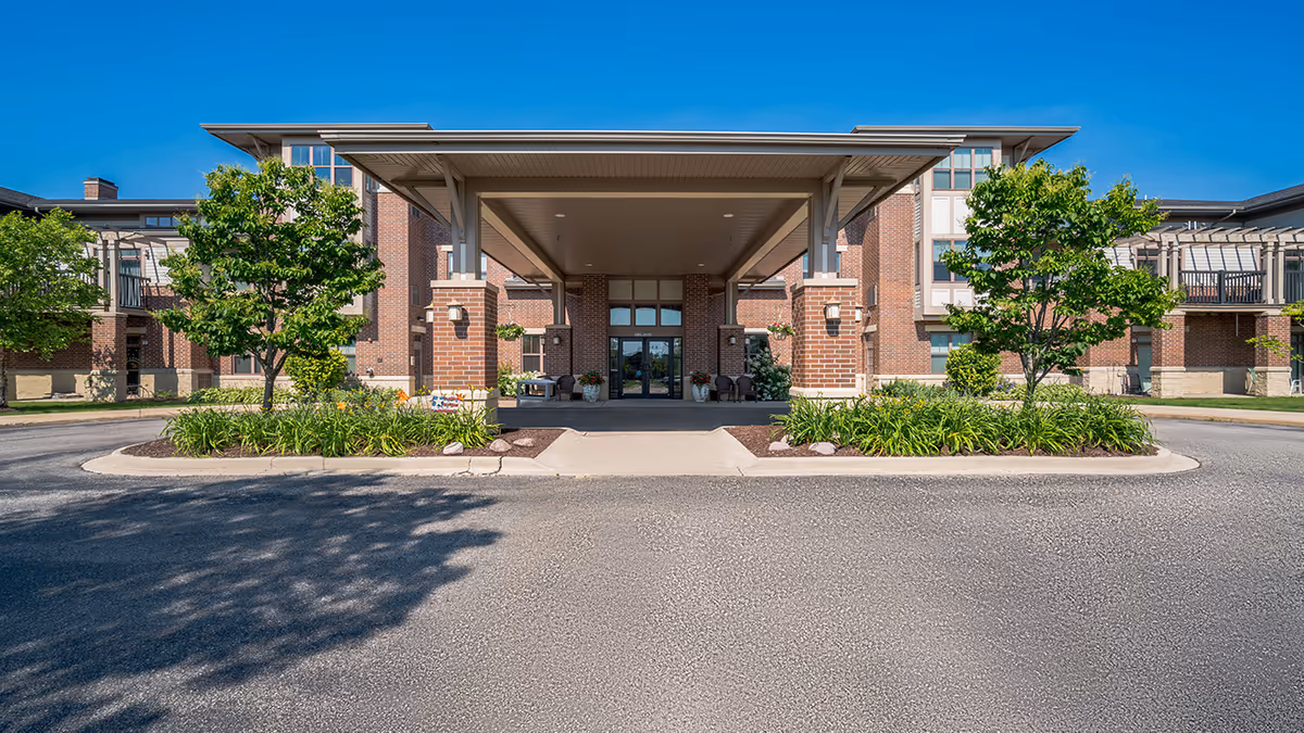 Front entrance of a senior living facility named Village at the Falls, featuring a covered drop-off area with brick pillars, landscaped greenery, and a clear blue sky.