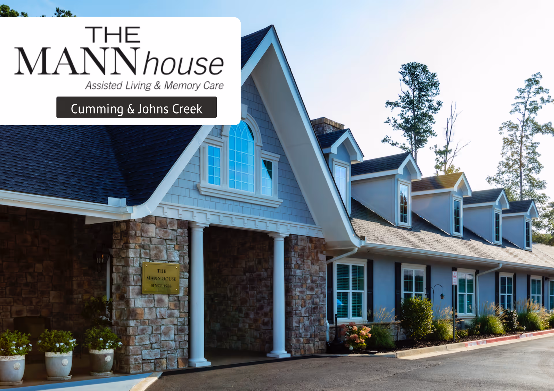 Exterior view of The Mann House assisted living and memory care facility in Cumming & Johns Creek, showing a building with stone and blue siding, white columns, multiple windows, and a paved driveway with landscaping.